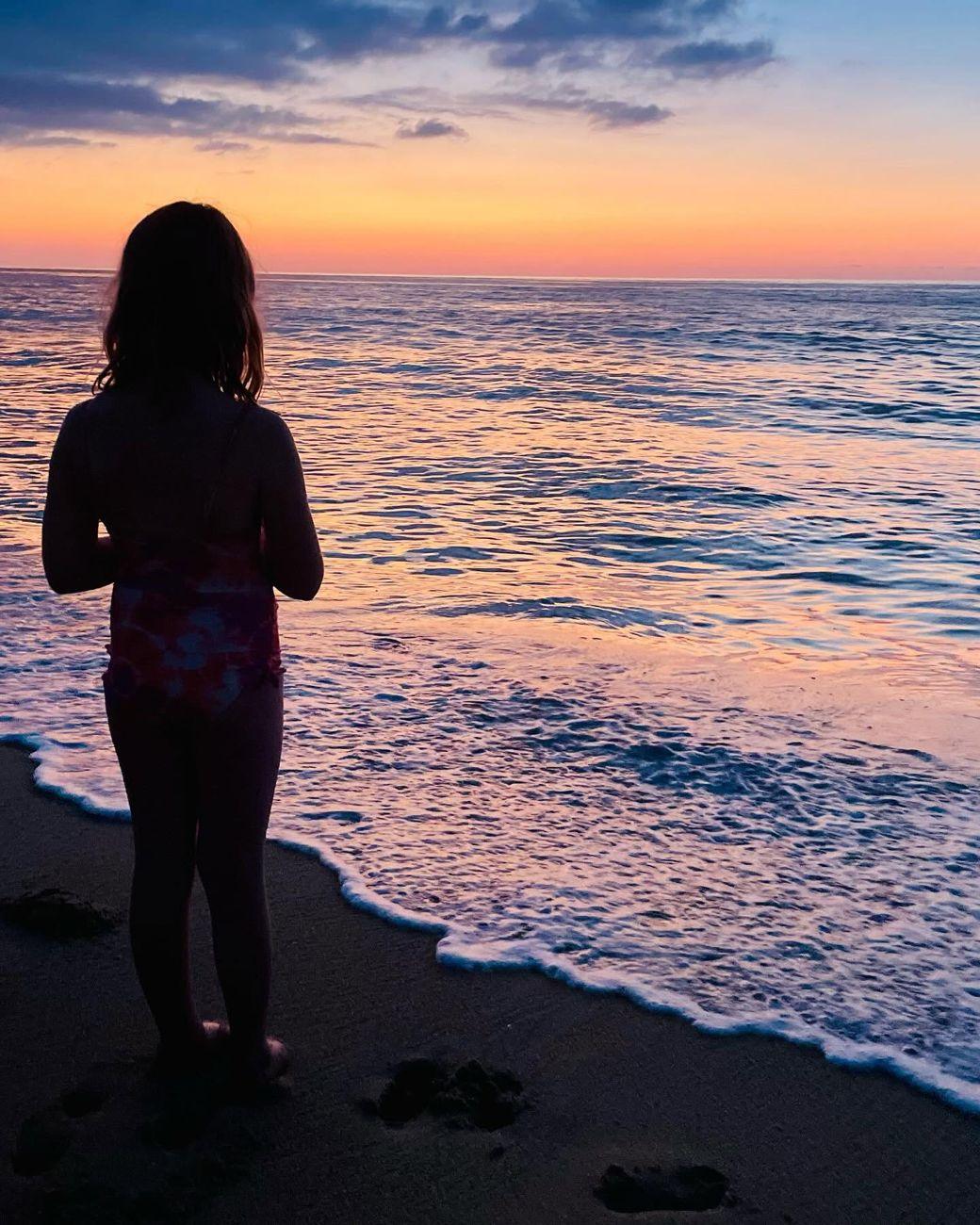 A child standing at the water’s edge, watching the sunset over the sea
