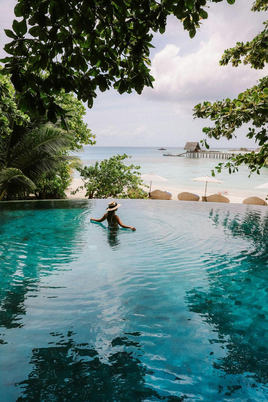 A woman in a sun hat resting in an infinity pool that looks out to a beach and pier